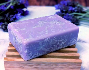 May include: A rectangular bar of light purple soap with a bubbly, foamy texture on its surface. The soap rests on a slatted wooden soap dish, with sprigs of purple lavender flowers visible in the background, creating a clean and natural aesthetic.