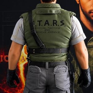 Könnte beinhalten: Eine Sammler-Actionfigur trägt ein hellgraues Kurzarmhemd und eine grüne taktische Weste mit dem aufgedruckten Text S.T.A.R.S. SPECIAL TACTICS AND RESCUE SERVICE auf dem Rücken. Die Figur trägt schwarze Handschuhe, einen schwarzen Gürtel und eine graue Hose.