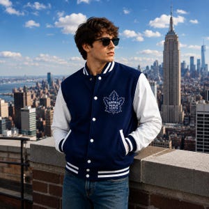 Op de afbeelding: Een marineblauw varsity-jack met witte mouwen, voorzien van een wit Toronto Maple Leafs-logo op de borst. Het jack heeft een sluiting met drukknopen, een geribbelde kraag en gestreepte boorden en zoom in marineblauw en wit.