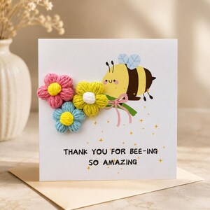 May include: A square greeting card with a white background featuring a cartoon bee illustration and three dimensional yarn flowers in pink, yellow, and blue. The text reads, THANK YOU FOR BEE-ING SO AMAZING, accompanied by small gold star accents.