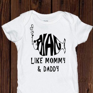 May include: White baby onesie with a black and white fish design. The fish is made up of the letters "DAD" and has bubbles coming out of its mouth. The text "LIKE MOMMY & DADDY" is printed below the fish.