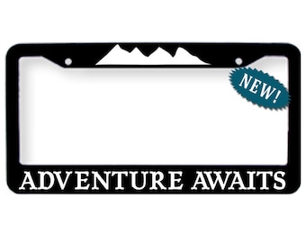 Op de afbeelding: Zwarte kentekenplaathouder met een wit berg silhouet en de tekst "ADVENTURE AWAITS". Een teal blauwe sticker met het woord "NEW!" zit op de houder.