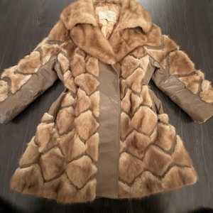 Puede incluir: Un abrigo color canela con un cuello grande y un patrón geométrico de parches de piel alternados con paneles de cuero liso. La prenda tiene mangas largas y un diseño estructurado con una etiqueta interior visible en la zona del cuello.
