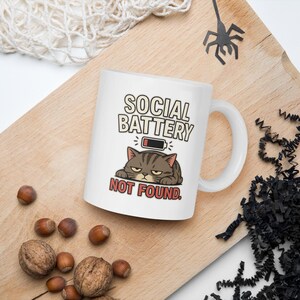 May include: A white ceramic mug featuring a graphic of a grumpy brown tabby cat lying down with a low battery icon above it. The text reads SOCIAL BATTERY in black letters and NOT FOUND in red letters below the cat illustration.