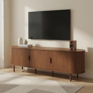 Op de afbeelding: Een lang, ovaalvormig houten mediameubel met verticaal latjeswerk op de kastdeuren en dunne, cilindrische poten. Een flatscreen-tv hangt aan de muur boven het meubel, met een kleine luidspreker en twee witte vazen op het blad.
