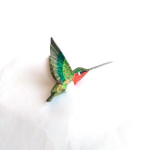 Puede incluir: Figurita de colibrí hecha a mano con las alas extendidas. El ave presenta una garganta roja, alas verdes y doradas y un pico largo y delgado. El cuerpo es una mezcla de verde y dorado, con detalles blancos.