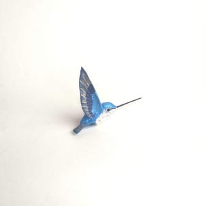 May include: A small, blue hummingbird figurine with outstretched wings. The bird has a long, black beak and white detailing on its chest. The wings are a mix of blue and gray, and the figurine appears to be made of wood.