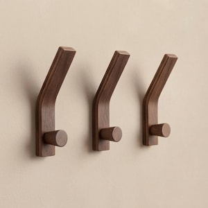 Könnte beinhalten: Drei dunkelbraune Wandhaken aus Holz, bestehend aus einer vertikalen Basis mit einem abgewinkelten oberen Arm und einem kleinen zylindrischen Stift, der aus dem unteren Bereich herausragt. Die Haken haben eine glatte Holzmaserung.