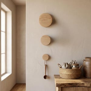 Könnte beinhalten: Drei kreisförmige Wandhaken aus hellem Holz, die vertikal montiert sind. Der untere Haken hält einen Holzlöffel an einer dünnen Schnur. Ein geflochtener Korb mit zwei kleinen Stoffbeuteln und einem Metallbehälter steht auf einer Holzoberfläche.