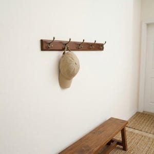 May include: A wall-mounted wooden coat rack with five bronze-toned metal hooks. A tan baseball cap hangs from the second hook from the left. The rack features a smooth, dark brown finish and is mounted horizontally on a plain white wall.