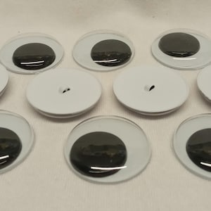 May include: A collection of ten round craft eyes, each featuring a clear plastic dome over a solid black pupil. Some of the eyes are positioned with the white plastic backing facing upward, revealing a small central hole for attachment.