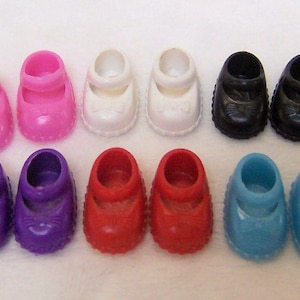 May include: A collection of ten miniature doll shoes arranged in two rows. The shoes are molded in pairs of pink, white, black, purple, red, and light blue. Each shoe features a rounded toe, a single strap across the top, and a small bow detail.
