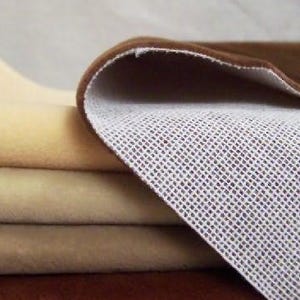 May include: A stack of folded fabric pieces in shades of cream, beige, and brown. The top brown piece is folded over to reveal a white, grid-like, textured backing. The fabric has a soft, smooth surface appearance with a consistent, dense weave.