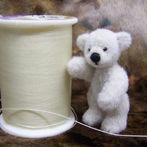 May include: A miniature white teddy bear with a black nose and dark eyes standing next to a large spool of off-white sewing thread. The bear has a fuzzy texture and is positioned to the right of the cylindrical spool, which features a white plastic base and top.