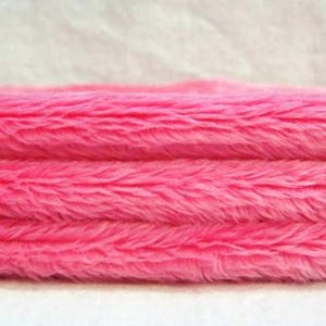 May include: A close-up view of a folded piece of bright pink faux fur fabric. The material features a dense, soft, and shaggy texture with long, synthetic fibers that create a layered appearance across the horizontal folds of the textile.