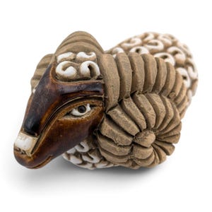 May include: A ceramic ram figurine featuring a dark brown glazed face with white accents on the eyes and nose. The ram has large, textured, tan-colored coiled horns and a body covered in a repeating pattern of white, rounded, raised shapes.