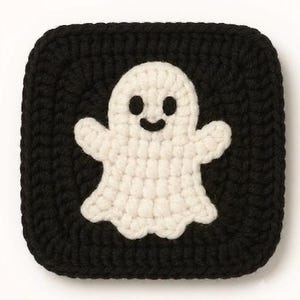 May include: A square crochet patch featuring a white ghost motif centred on a solid black background. The ghost has two small black circular eyes and a curved black mouth, all worked in yarn with a visible textured stitch pattern throughout.