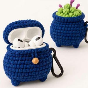 Puede incluir: Dos fundas de ganchillo azul oscuro para estuches de auriculares inalámbricos, con forma de pequeños calderos de tres patas. Una funda está abierta, mostrando auriculares blancos y un cierre de cuenta de madera. Ambas incluyen un mosquetón negro.