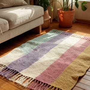 May include: A rectangular woven rug featuring wide horizontal stripes in alternating colors of light green, lavender, cream, dusty rose, and mustard yellow. The rug has a textured knit appearance and includes long fringe tassels on both ends.