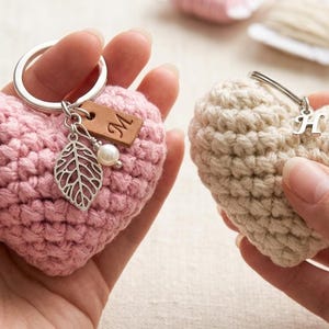 Op de afbeelding: Twee gehaakte sleutelhangers in de vorm van een hart. Het roze hart heeft een zilveren ring met een bruin leren label met de letter M, een zilveren blaadje en een witte kraal. Het beige hart heeft een zilveren ring met een zilveren letter H-bedel.