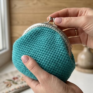 May include: A teal crochet coin purse featuring a textured, grid-like stitch pattern. The pouch is secured with a silver-toned metal kiss-lock frame that has an intricate, embossed decorative pattern along the outer edge of the closure.