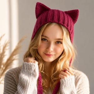 Pode incluir: Um gorro de malha magenta profundo com duas formas proeminentes e pontiagudas de orelhas de gato no topo. O gorro é construído com uma textura canelada vertical e inclui dois longos laços de malha a condizer que pendem dos lados.