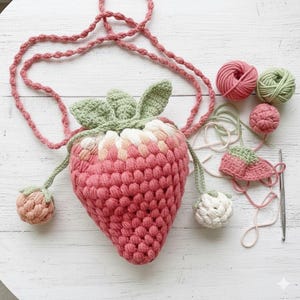 May include: A strawberry-shaped crochet bag with a long pink strap, featuring a textured pink and white body and green leaves at the top. Two small crochet strawberry charms hang from the sides, accompanied by yarn balls and a metal crochet hook.