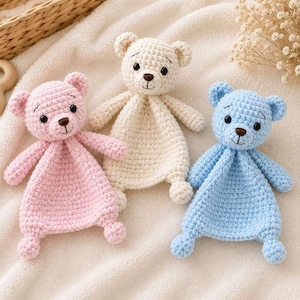 May include: Three crochet security blankets featuring bear heads with embroidered eyes, noses, and mouths. The bears are in soft pastel colors: one light pink, one cream, and one light blue. Each has a flat, square-shaped crocheted body.