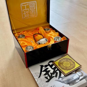 May include: A black box with a gold interior lining containing a silver teapot with a braided handle and four small silver cups. The box lid features a white logo. Beside the box lies a booklet with Chinese characters and a small square card.