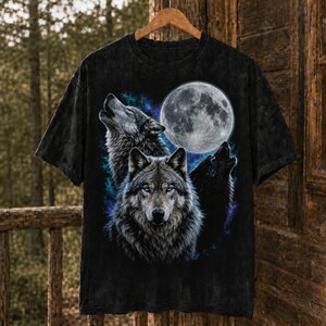 May include: A black short-sleeve t-shirt featuring a graphic print of three wolves and a full moon. The central wolf faces forward with blue eyes, while two other wolves are shown in profile howling against a backdrop of blue nebula clouds.