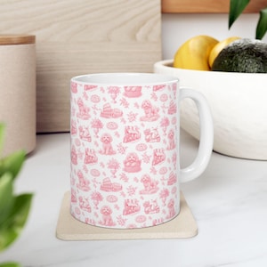 May include: A white ceramic mug featuring a repeating pink toile-style pattern. The design includes illustrations of small dogs, scooters, the Colosseum, floral arrangements, and baskets, all rendered in a monochromatic pink ink on a white background.