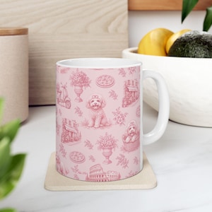 May include: A white ceramic mug featuring a repeating toile-style pattern in pink. The design includes illustrations of poodles, scooters, floral arrangements in urns, pizzas, Italian-style buildings, and the Colosseum on a soft pink background.