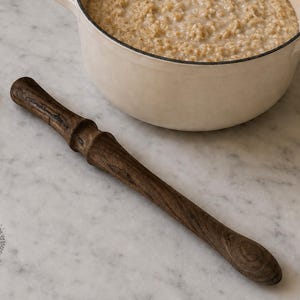 Può includere: Un mescolatore per porridge in legno marrone scuro con un manico tornito liscio e un corpo leggermente affusolato. L'utensile presenta una venatura distinta e un'estremità arrotondata, posizionato vicino a una pentola in ceramica bianca.