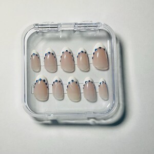 Peut inclure: Un ensemble de dix faux ongles avec une base nude et des pointes blanches façon French manucure, chacun orné d'un petit strass bleu au centre de la ligne de sourire. Les ongles sont disposés en deux rangées dans un boîtier de rangement carré.