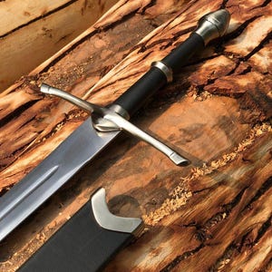 Op de afbeelding: Een stalen zwaard met een zwart handvat en een zilveren pommel, voorzien van een pareerstang en een centrale groef langs het blad. Naast het zwaard ligt een zwarte schede met een zilveren mondstuk en een rood lederen accent.