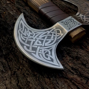 May include: A double-bitted axe featuring a polished metal head with intricate dark grey Celtic knotwork engravings. The handle is crafted from light-colored wood with a distinct grain, wrapped in dark brown leather strips for a secure grip.