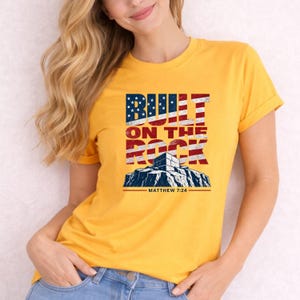 Könnte beinhalten: Ein leuchtend gelbes Kurzarm-T-Shirt mit einem Grafikdruck, der den Text BUILT ON THE ROCK und MATTHEW 7:24 zeigt. Das Design enthält eine stilisierte Abbildung einer Felsformation und ein Flaggenmuster in den Farben Rot, Weiß und Blau.