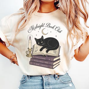 Può includere: Una t-shirt color crema con la grafica di un gatto nero sdraiato su un libro aperto sopra una pila di tre libri intitolati Spells & Charms, Potions & Brews e The Book of Shadows, con una candela e il testo Midnight Book Club sopra.