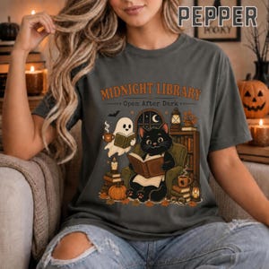 Può includere: Una t-shirt grigio scuro con una stampa grafica che riporta il testo "MIDNIGHT LIBRARY Open After Dark". L'illustrazione mostra un gatto nero e un fantasma bianco che leggono libri in una stanza con una libreria, una zucca e delle lanterne.