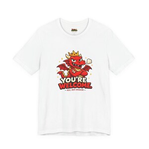 Könnte beinhalten: Ein weißes kurzärmeliges T-Shirt mit einer Grafik eines roten Drachen, der eine goldene Krone trägt und die Arme verschränkt hat. Der Text lautet YOU'RE WELCOME und REX, RED DRAGON. Der Drache hat kleine Flügel und Rauch aus der Nase.