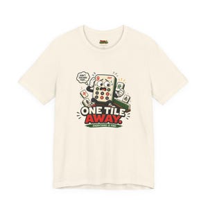 May include: A cream-colored t-shirt featuring a graphic of a stressed mahjong tile character with the text "DON'T DISCARD THAT...", "ONE TILE AWAY.", and "EVERYTHING IS FINE." The design includes other mahjong tiles scattered around the character.