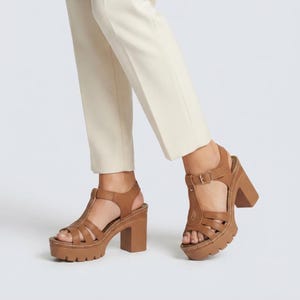 May include: A pair of tan leather platform sandals with a T-strap design, featuring a thick block heel, a lugged sole, and an adjustable ankle strap with a metal buckle. The sandals are worn with light-colored trousers, showcasing the open-toe style.