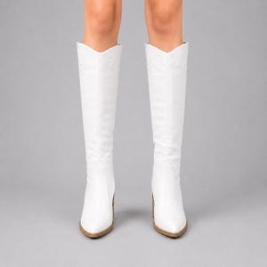 Puede incluir: Un par de botas blancas hasta la rodilla con punta afilada, tacón de bloque y una abertura en forma de V. Las botas presentan sutiles costuras tonales a lo largo de la caña y el pie, ofreciendo una apariencia blanca sólida y limpia de frente.