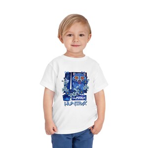 May include: White toddler t-shirt featuring a blue cartoon cat with large orange eyes, surrounded by blue floral accents. The shirt has the word "HUNTRA" in a stylized font. The design is centered on the front of the shirt.