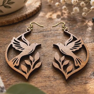May include: A pair of dark brown wooden drop earrings featuring a laser-cut design of a bird in flight above a leafy branch. The earrings are suspended from gold-toned metal hooks and have a smooth, flat finish with visible wood grain details.
