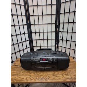 May include: A black RCA portable boombox featuring a top-loading CD player, a digital display with a red light, integrated speakers on both sides, a carrying handle, and a long silver telescopic antenna extending from the back right side.