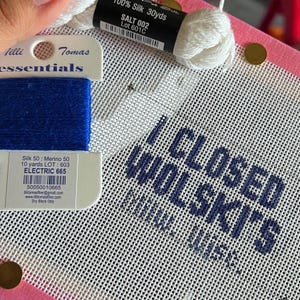 May include: A white needlepoint canvas featuring the dark blue stitched text I CLOSED WOLSKI'S, accompanied by a skein of white silk thread labeled SALT 002 and a card of blue silk and merino blend thread labeled ELECTRIC 665 with a 30-yard length.