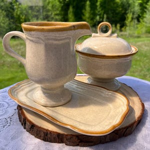 Peut inclure: Un ensemble composé d'un pot à lait, d'un sucrier avec couvercle et d'un plateau rectangulaire assorti. Les pièces sont blanc cassé avec une texture mouchetée et un bord brun distinct. Chaque élément possède une base sur pied et un bord festonné.