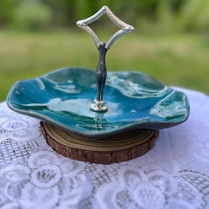Peut inclure: Un plateau de service à un étage composé d'une coupelle en céramique bleu sarcelle aux bords ondulés et à la finition brillante. Une poignée centrale en métal argenté avec une boucle en forme de losange surmonte le centre du plat.