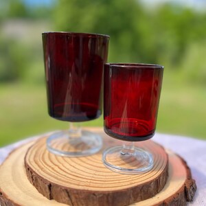 Peut inclure: Deux verres à pied en verre rouge profond avec des tiges et des bases circulaires transparentes. Le verre le plus grand est placé derrière le plus petit, tous deux dotés de bols cylindriques lisses et d'une finition translucide.